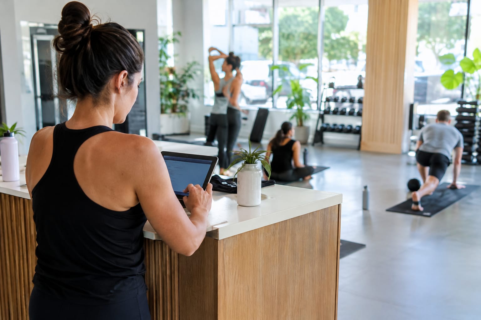 Fitness studio owner checking a tablet at reception while members stretch on mats in the background