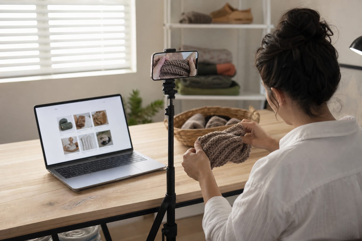 Handmade-goods seller photographing a product with a laptop showing a shop listing behind
