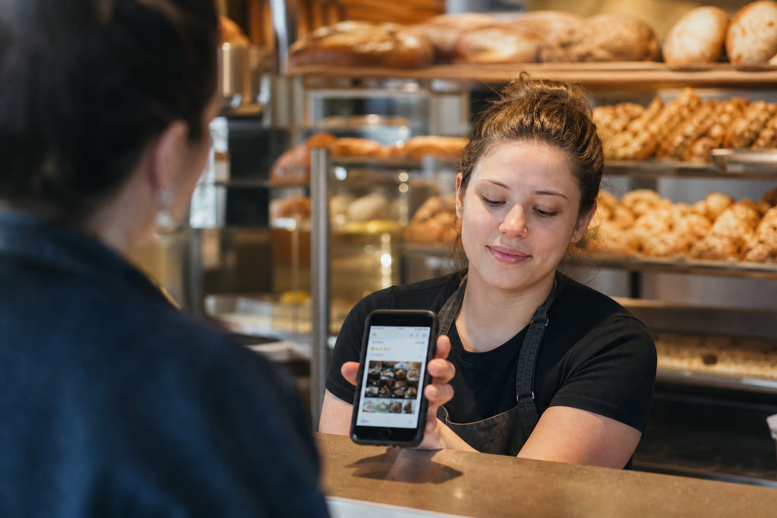 Bakery owner holding a phone showing her Google Business Profile with reviews, pastries behind her.