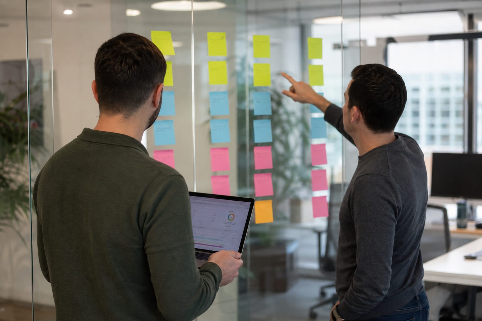 Two colleagues at a glass wall of grouped sticky notes, one holding a laptop with a site audit dashboard.