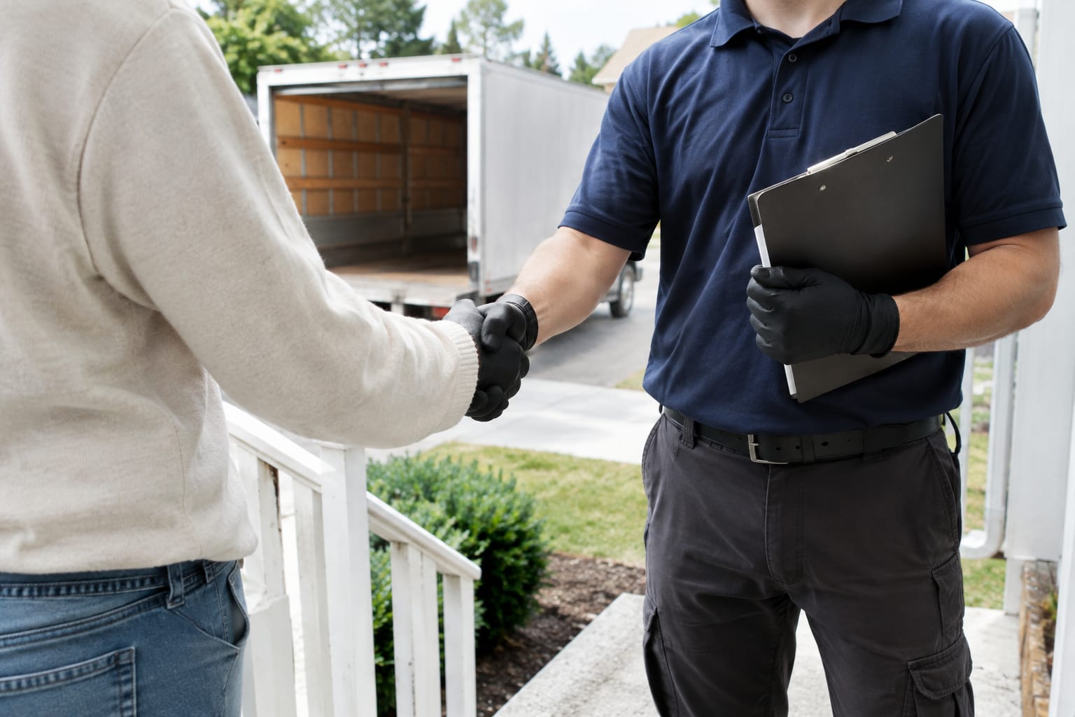Homeowner shaking hands with a mover holding a clipboard on a front porch, empty moving truck behind them
