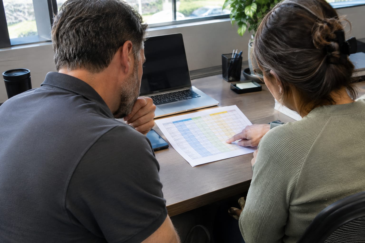 Two people at a desk pointing at one row of a printed four-column comparison spreadsheet