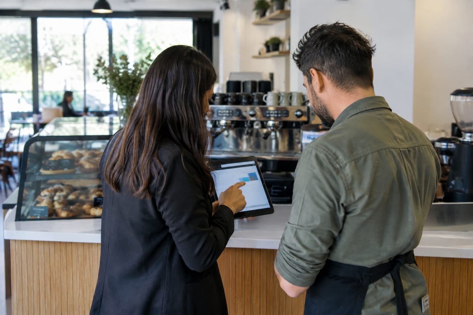 Account manager showing a café owner a tablet beside an espresso machine and pastry case