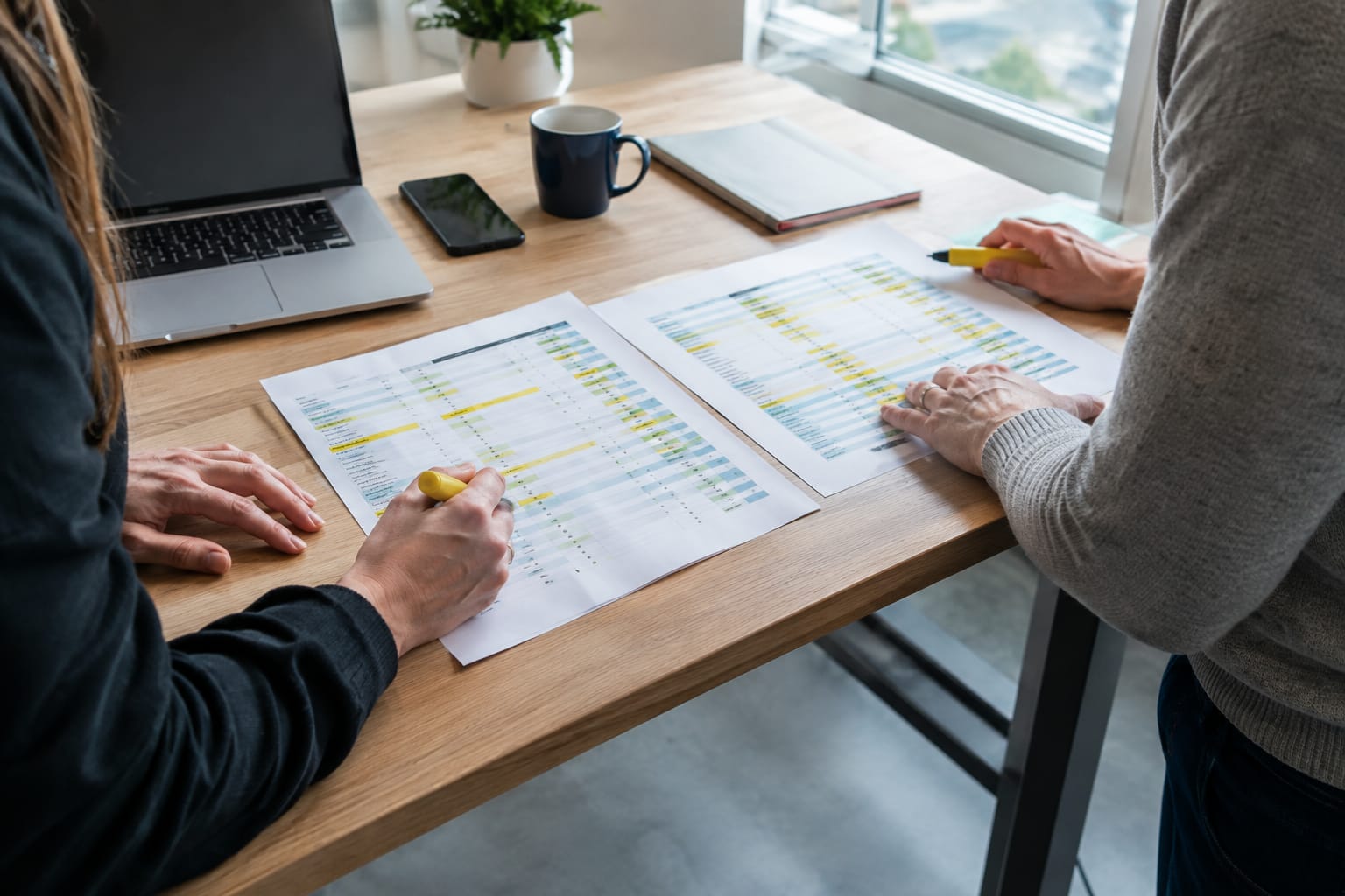 Two partners at a standing desk highlighting rows on printed comparison sheets with a yellow marker