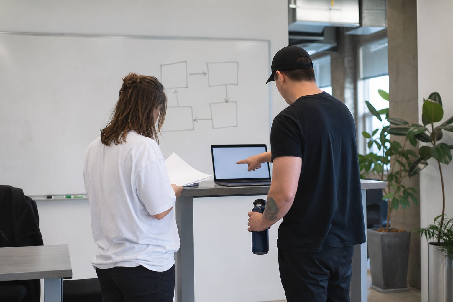 Two colleagues reviewing a draft at a standing desk with a four-box workflow on the whiteboard