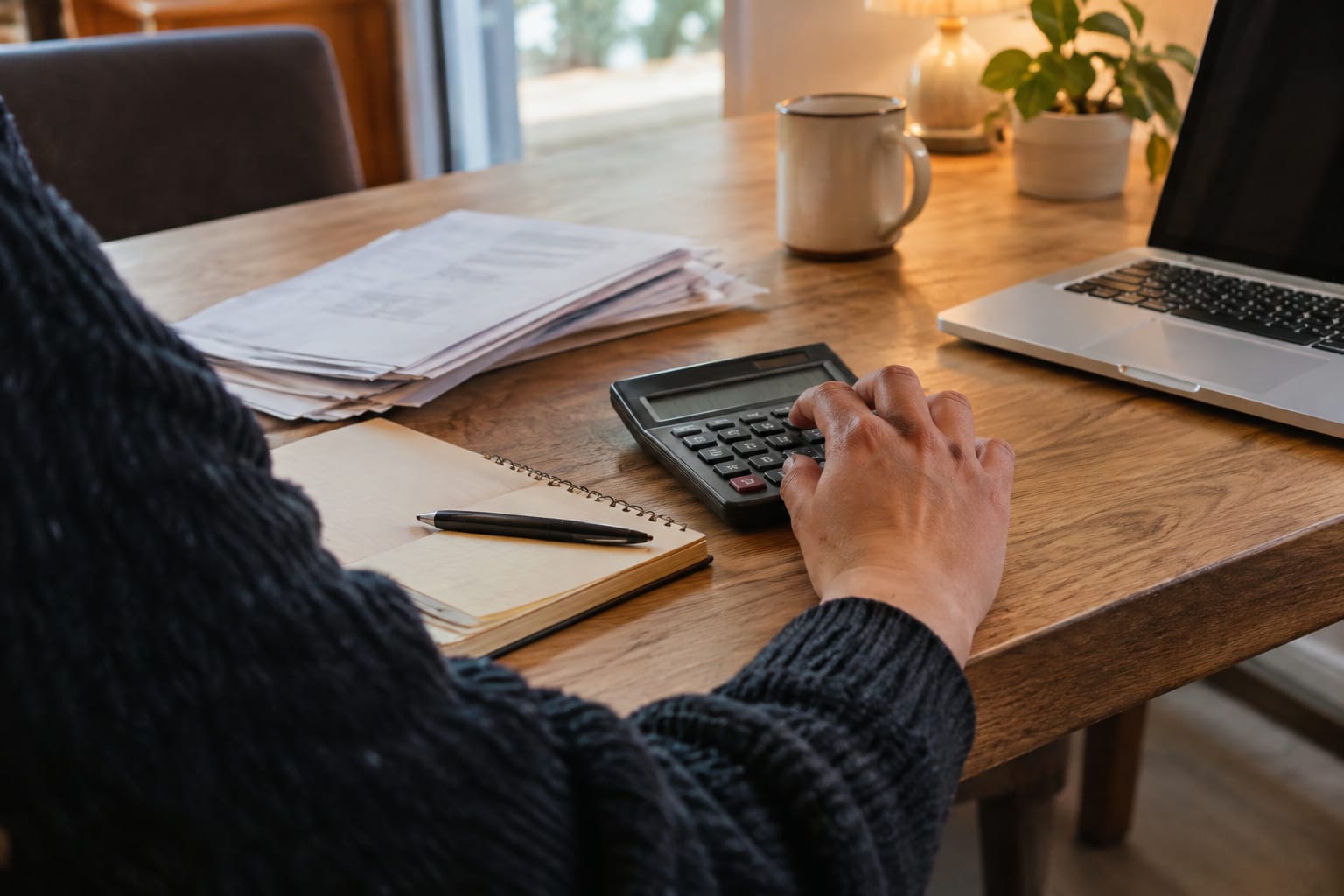 Agency owner calculating per-client margins with a calculator, printed invoices and a handwritten notebook