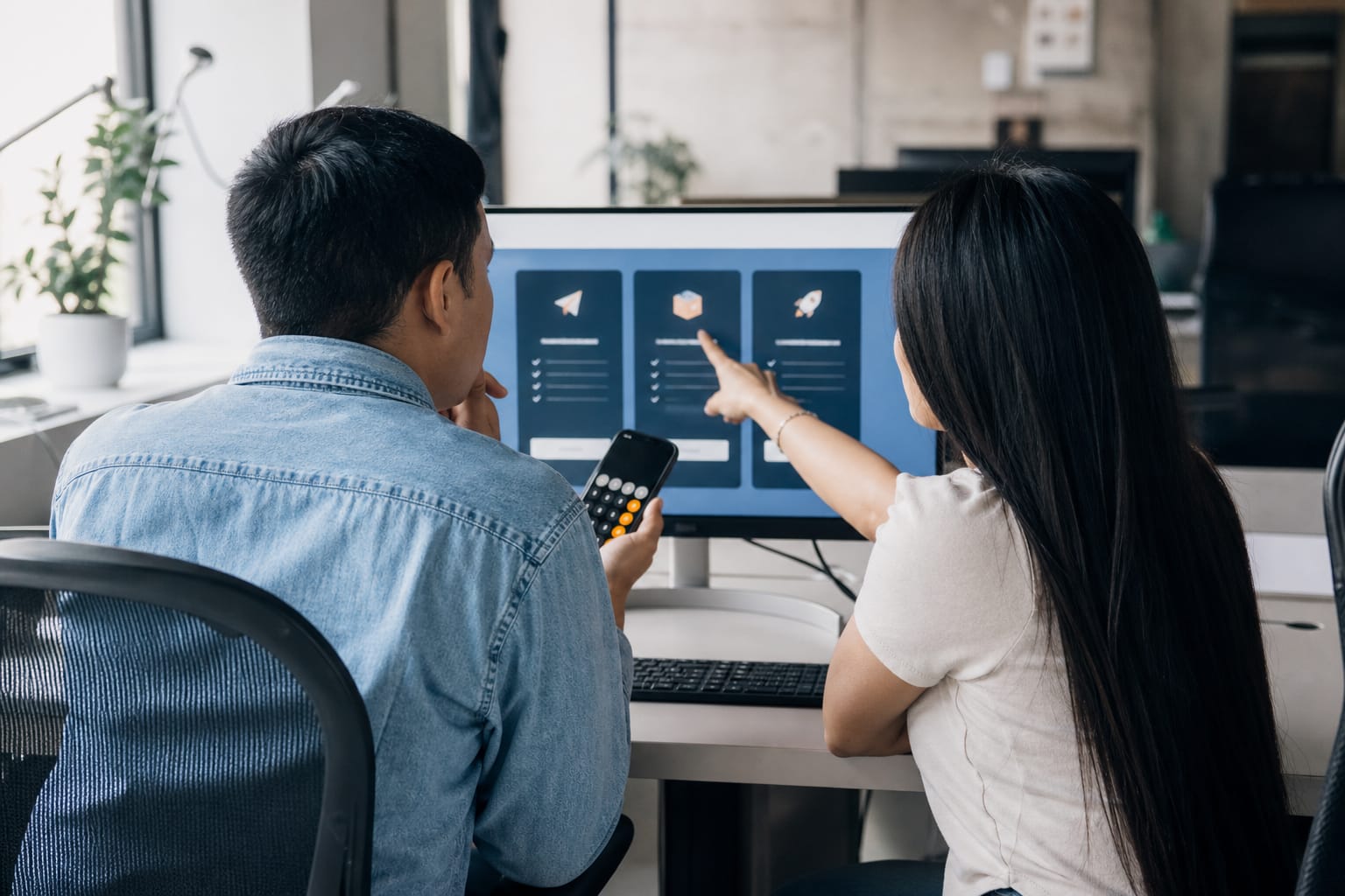 Two marketers comparing subscription plan cards on screen while one holds a calculator app