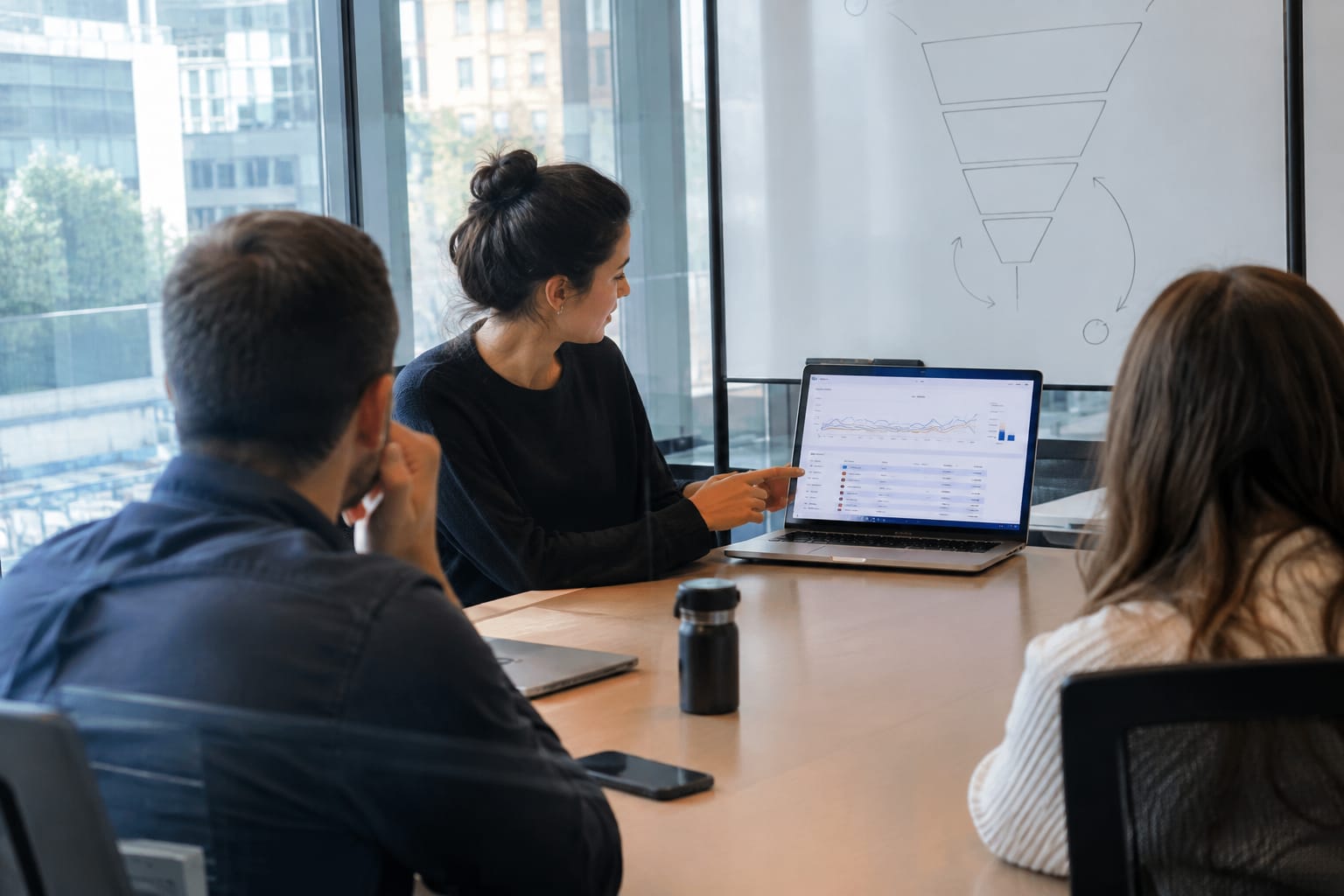 Manager showing two colleagues a laptop dashboard of weekly AI citations, with a funnel sketch on a whiteboard